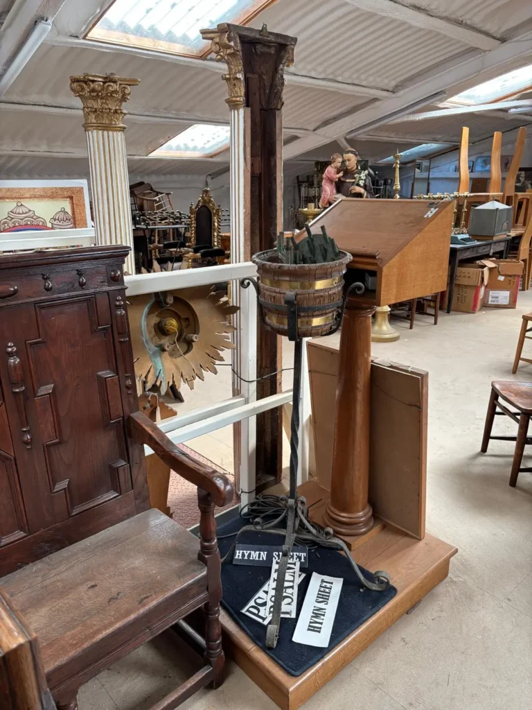 A carved oak armchair beside a Corinthian column, a gilded sunburst, a brass pot on an iron stand and a hymn board