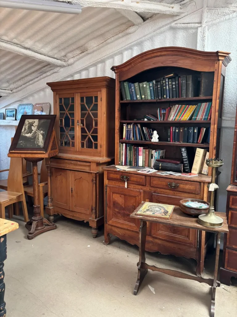 An antique pine bookcase full of old books beside a glazed cabinet, with a framed print displayed on a reading stand