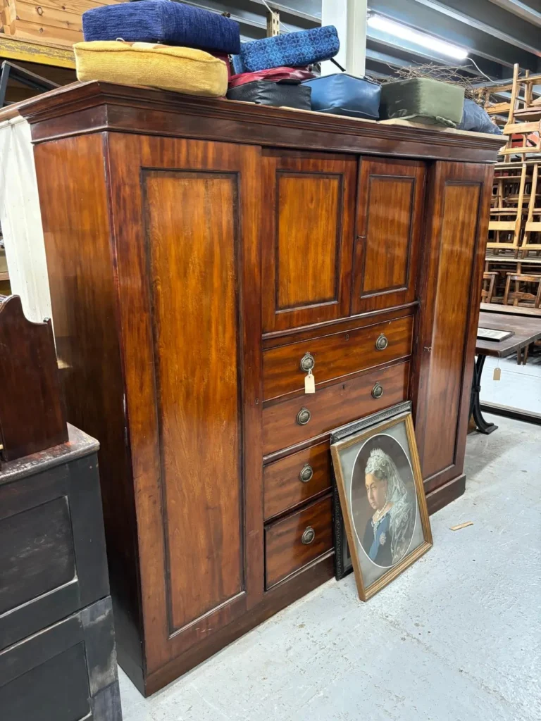 A tall mahogany cupboard with drawers, topped with a stack of colourful church kneelers, with a framed portrait leaning against it