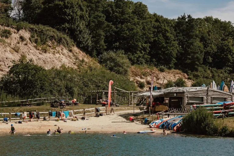 The sandy beach at Buckland Park Lake, with swimmers in the water, paddleboards stacked by the boathouse and wooded hills behind