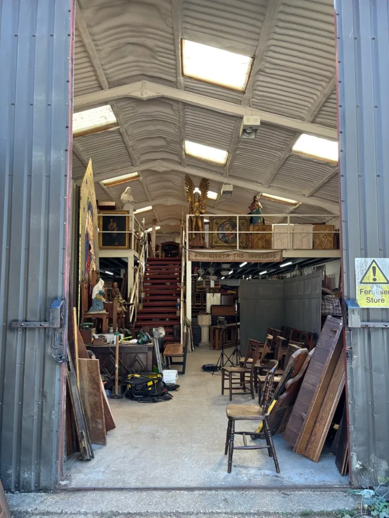 The warehouse doors open onto aisles of church furniture, with a red staircase leading up to the mezzanine